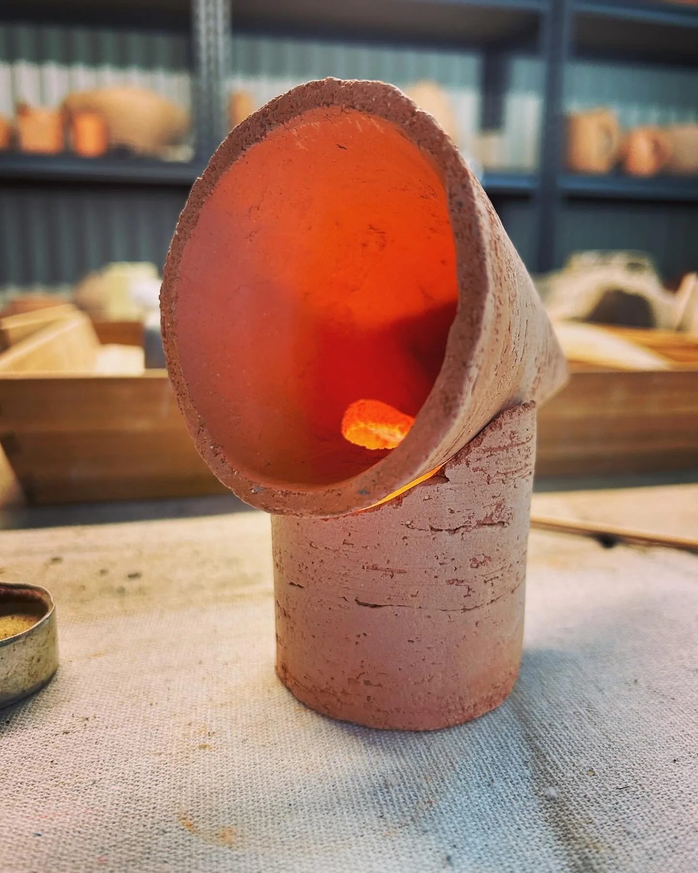 Close-up of a conical ceramic sculpture with an open circular top, resting on a textured surface. The sculpture has a rough, matte finish and appears pinkish in color. The interior of the opening is glowing orange, possibly indicating a light source 