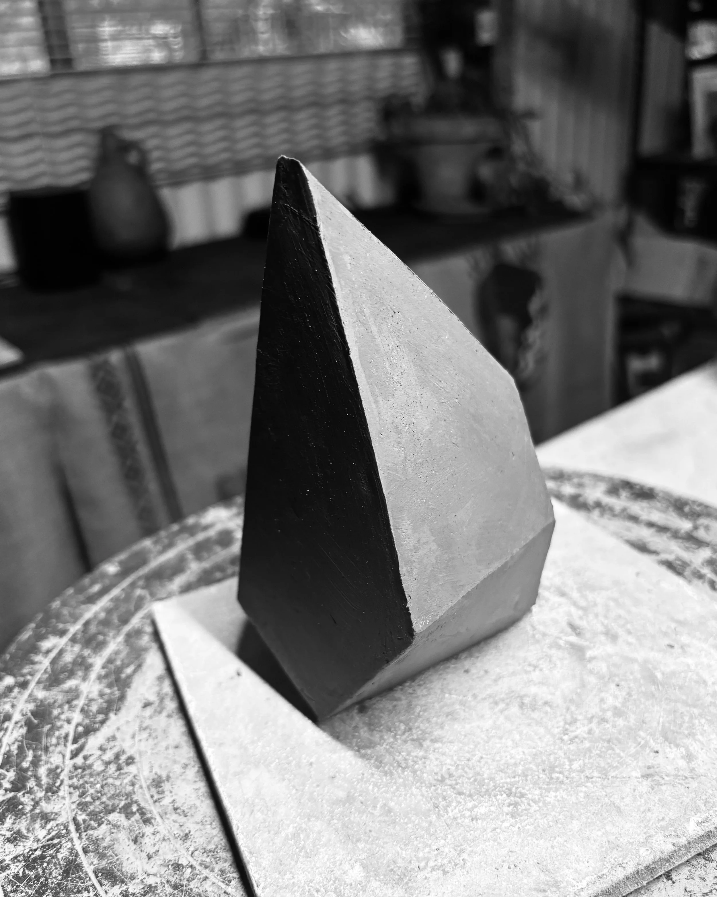 A black and white photograph of a pyramid-shaped object, possibly made of stone, placed on a dusty surface in a workshop or studio setting.