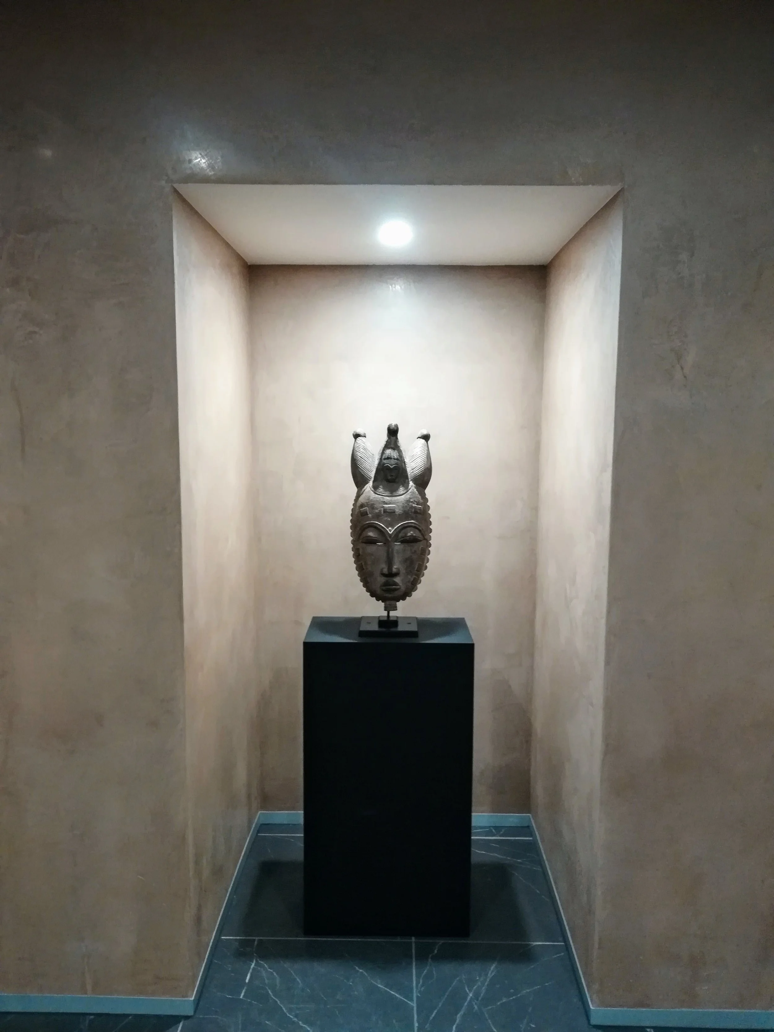 A sculpture of a face with a headdress, placed on a black pedestal inside a recessed display with spotlighting.