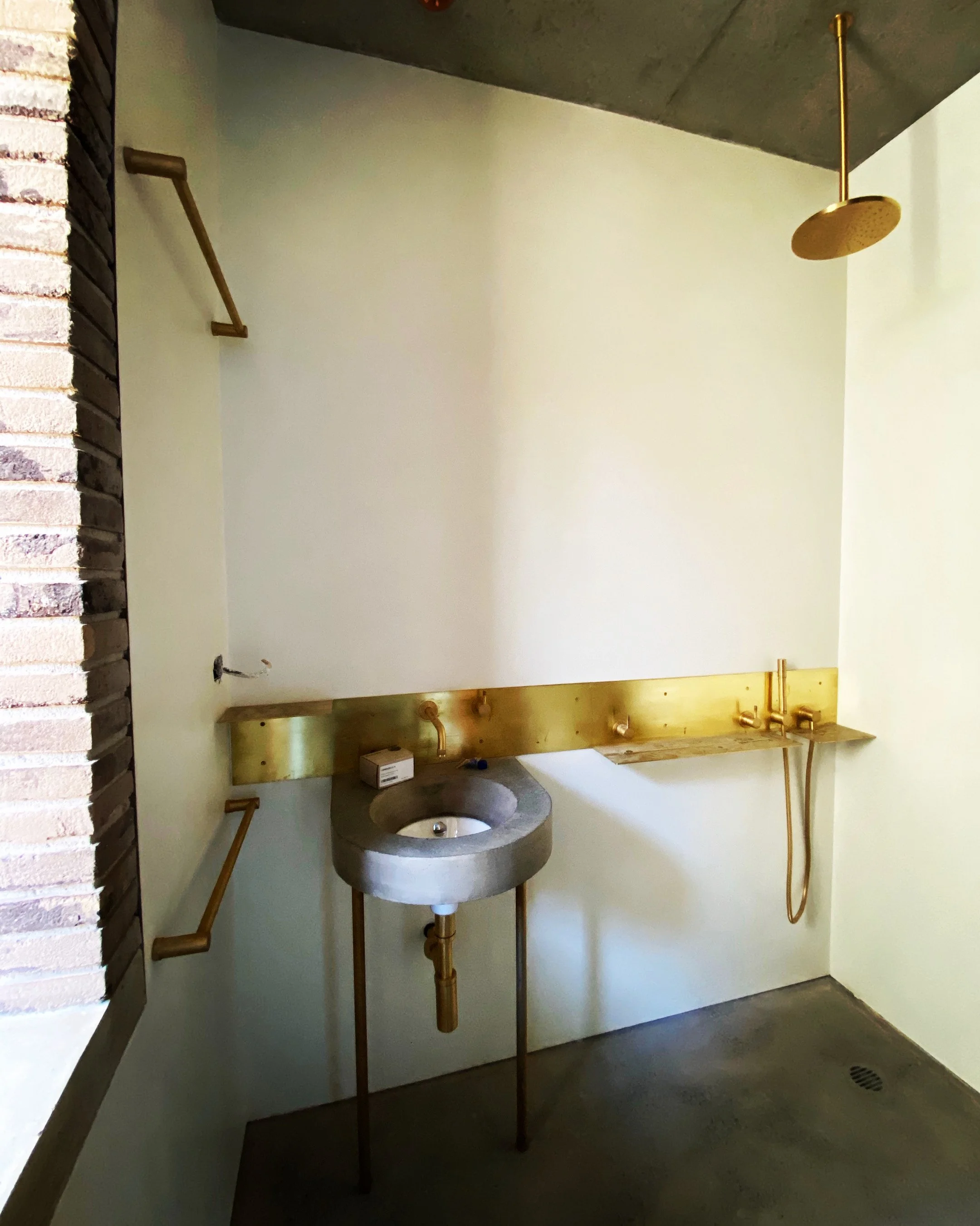 Minimalist bathroom with a concrete sink, gold fixtures, a rain showerhead, and a gold shelf on a white wall.
