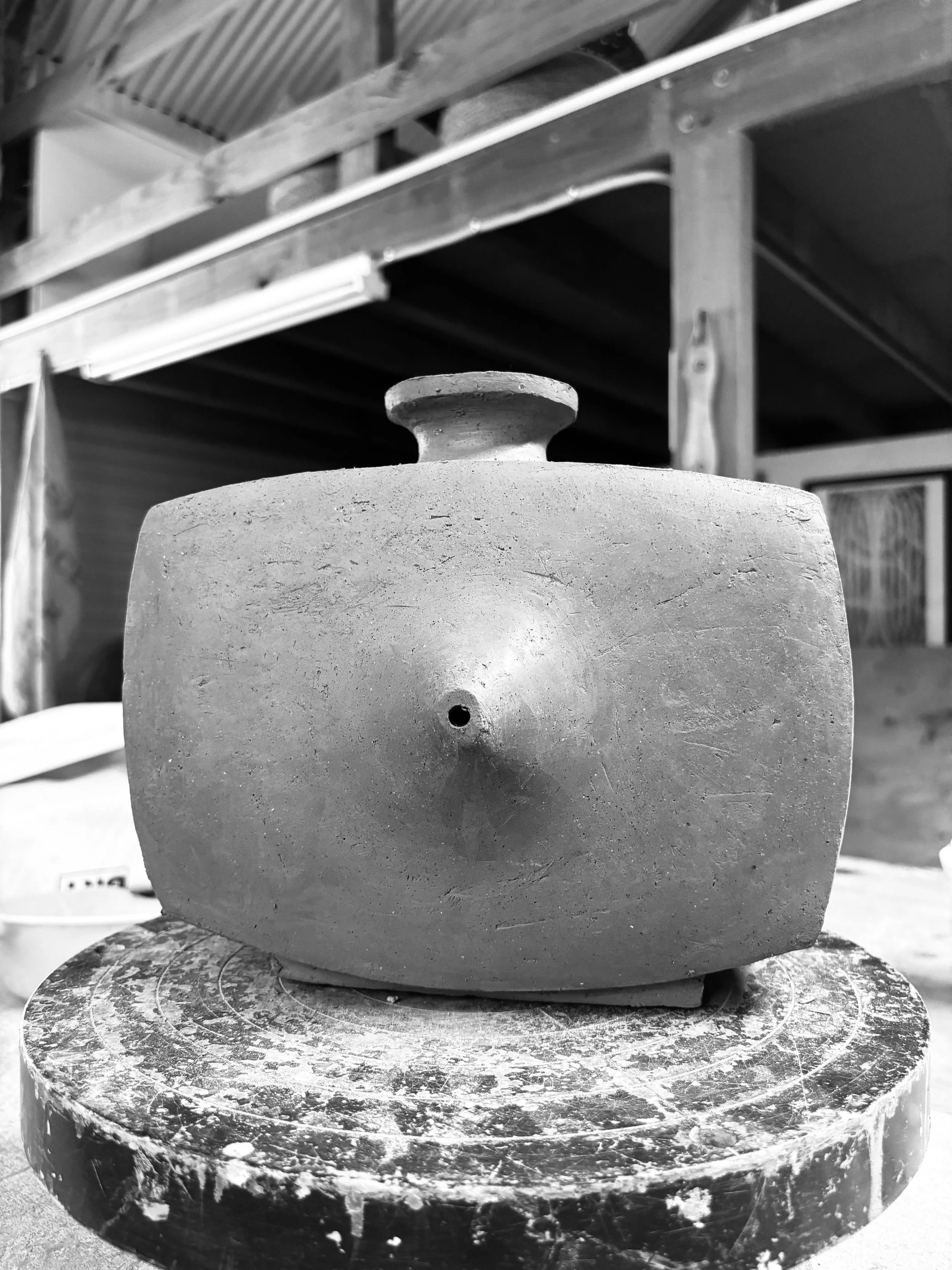 A clay pottery vessel with an opening on its face, placed on a round, dusty shelf in a workshop.