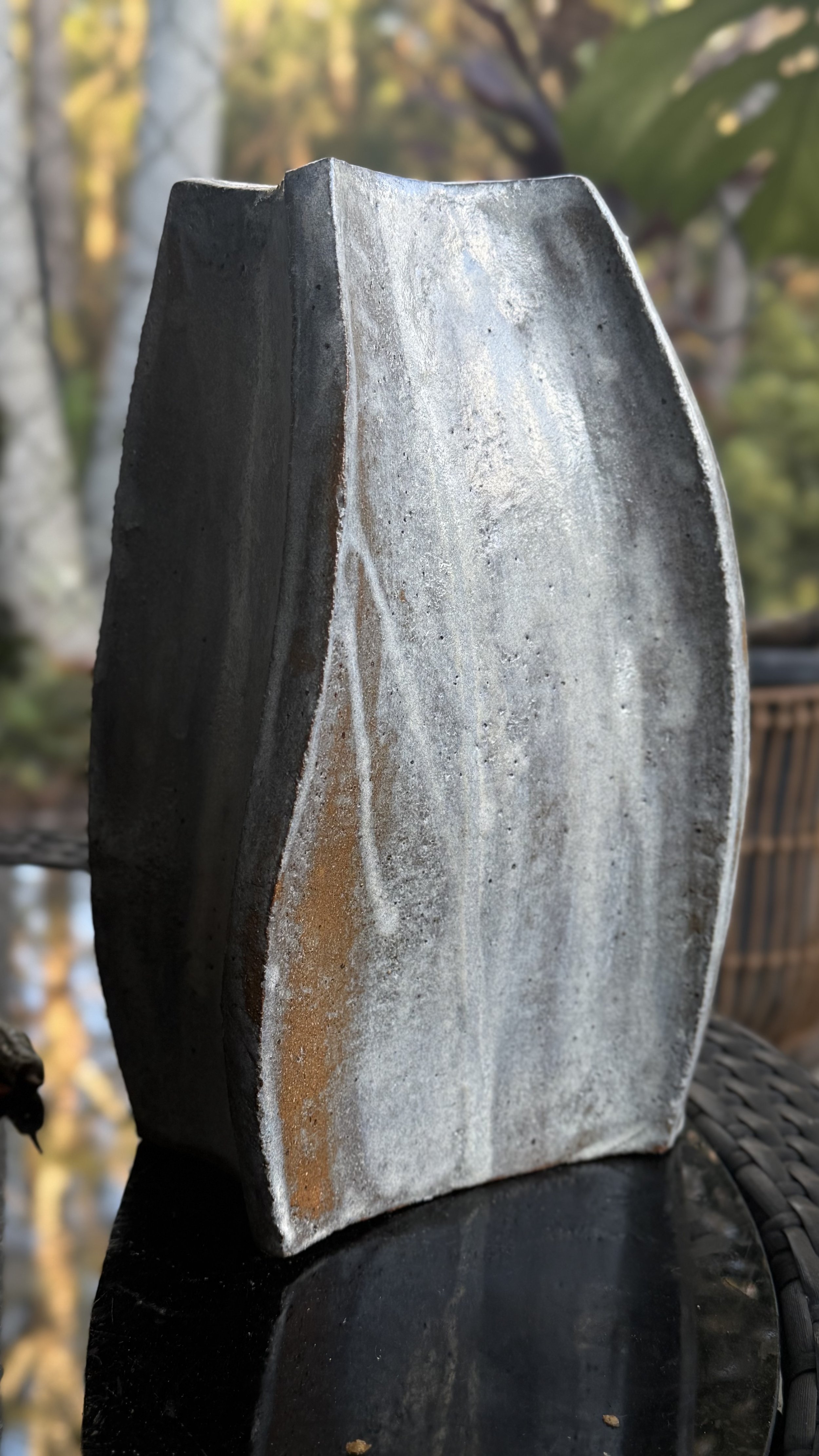 Close-up of a large, gray, polished stone sculpture with a smooth, curved surface, placed outdoors with blurred trees and foliage in the background.