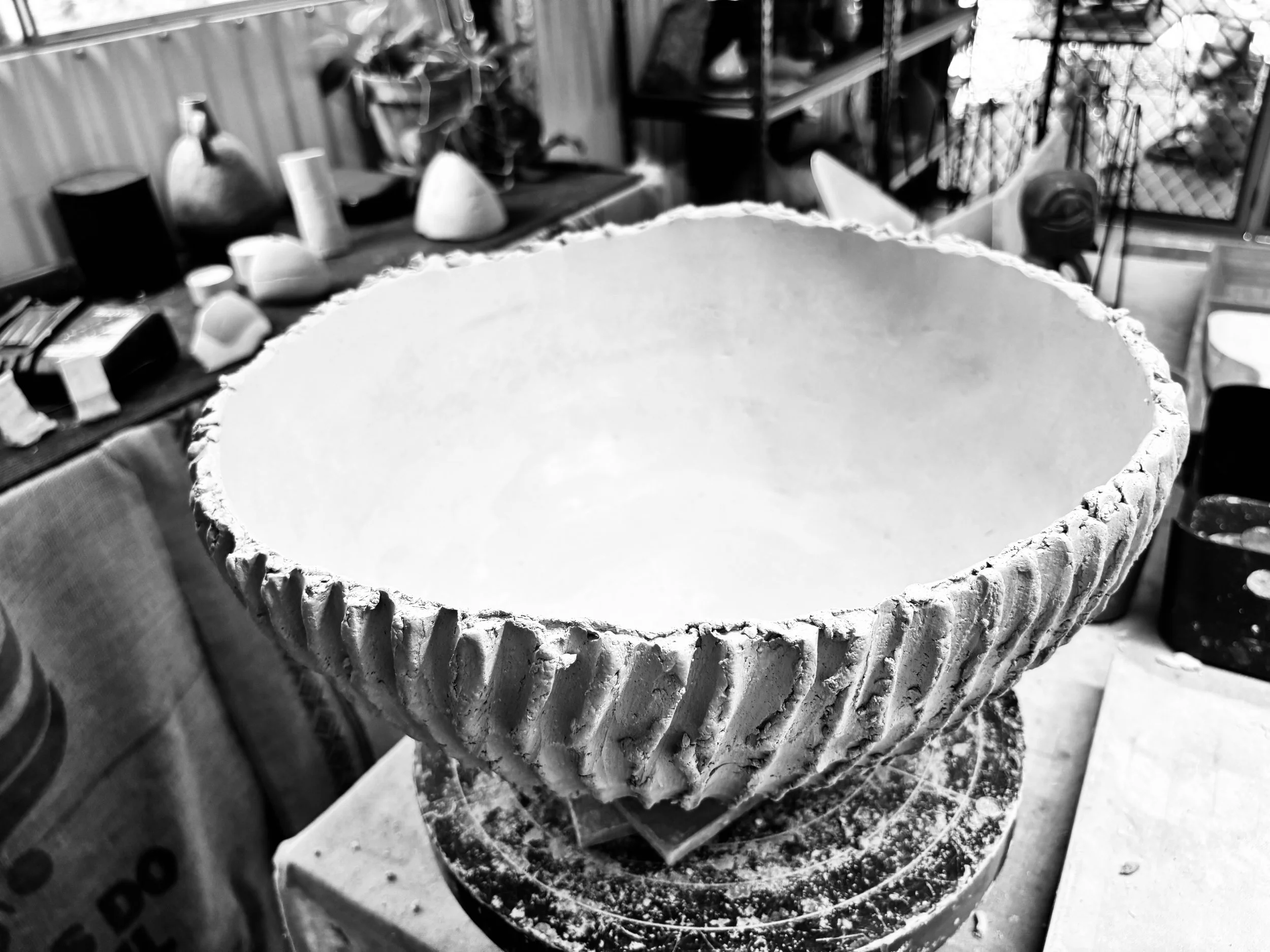 A large, empty, intricately carved bowl or vessel on a stand in a workshop or craft room, surrounded by various objects and tools.