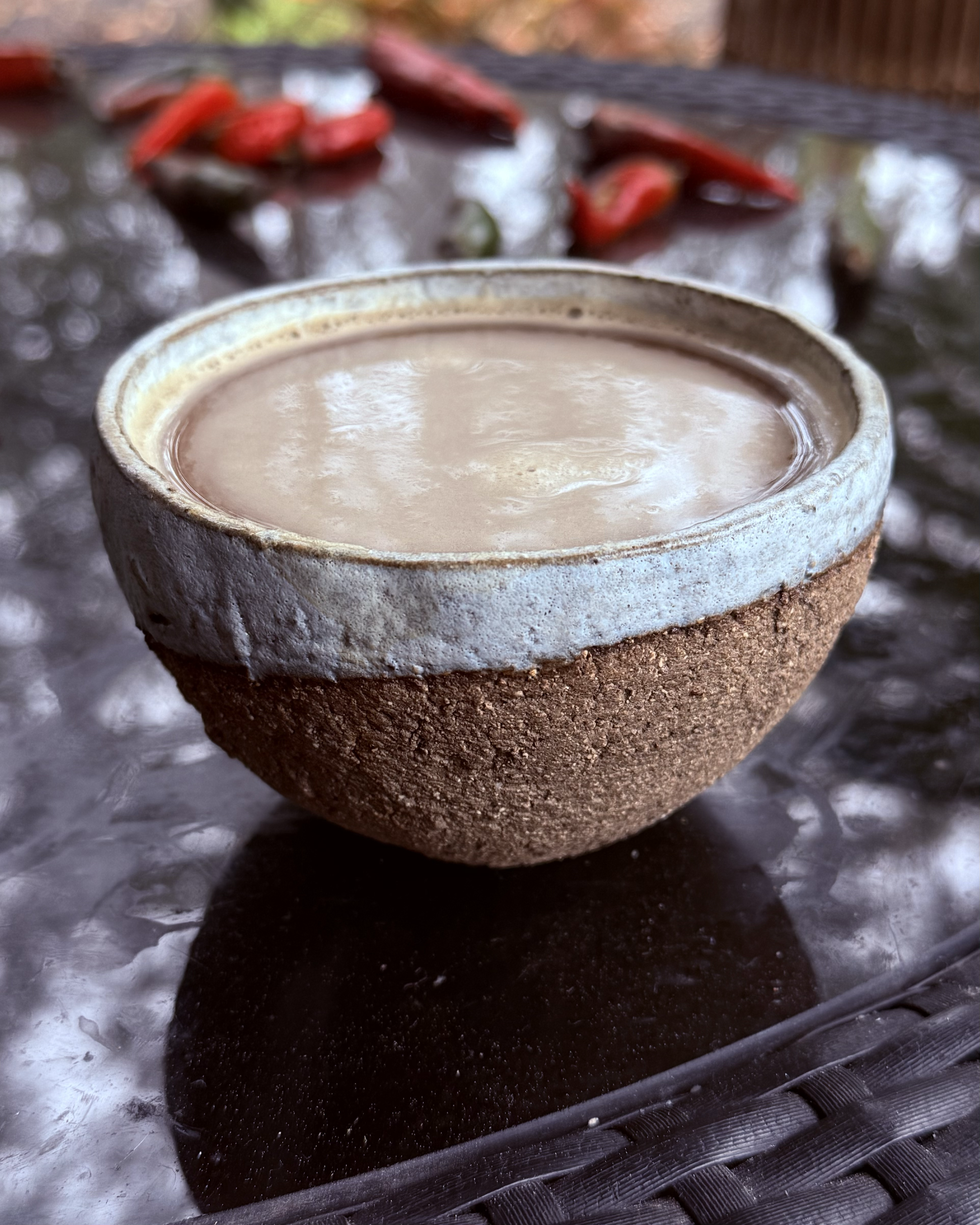 A ceramic bowl filled with a light brown beverage on a black textured surface, with blurred red chili peppers and greenery in the background.