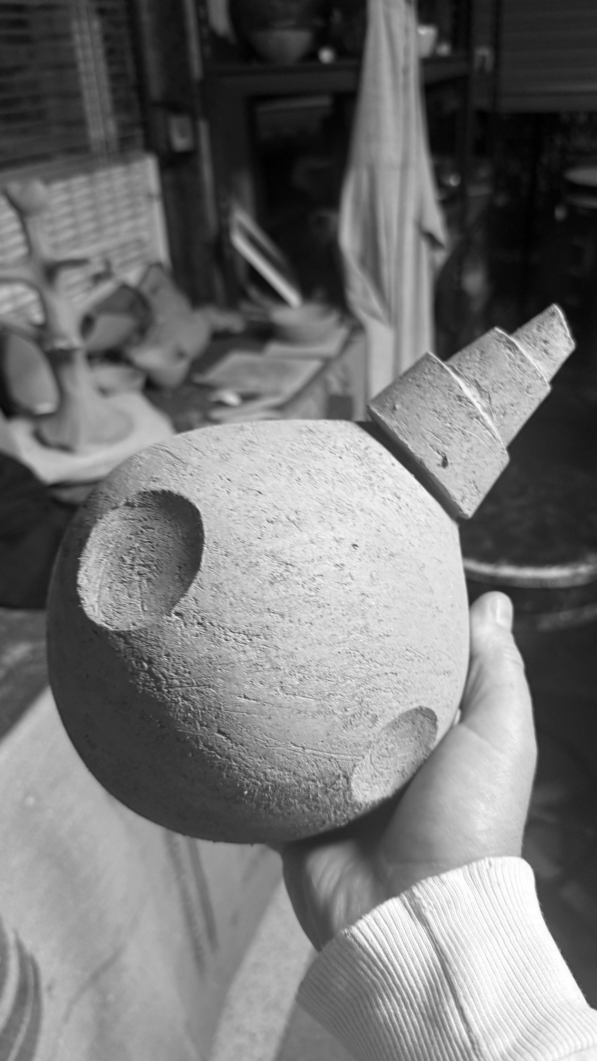 A hand holding a textured round ceramic object with two rectangular protrusions on top, in a workshop setting with shelves and various ceramic items in the background.