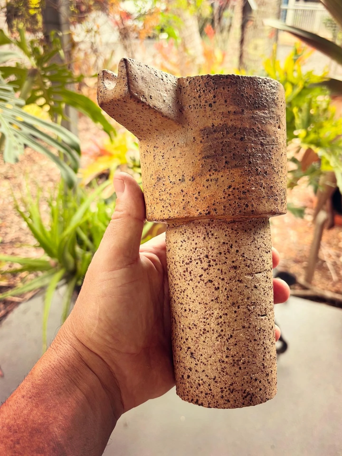 A person's hand holding a large, textured, two-part ceramic or stone sculpture outdoors, with green plants and trees in the background.