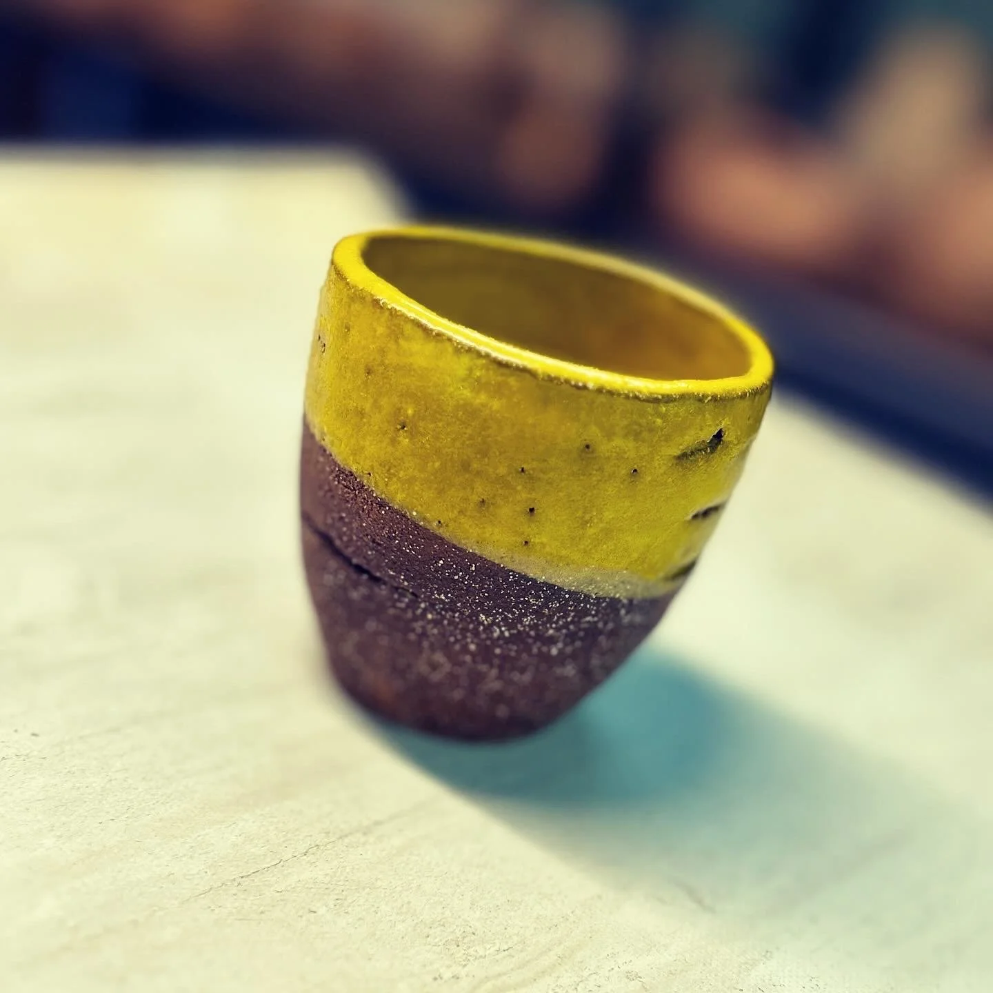 A two-tone ceramic cup with a yellow upper half and a dark brown lower half, placed on a light-colored surface, with a blurred background.