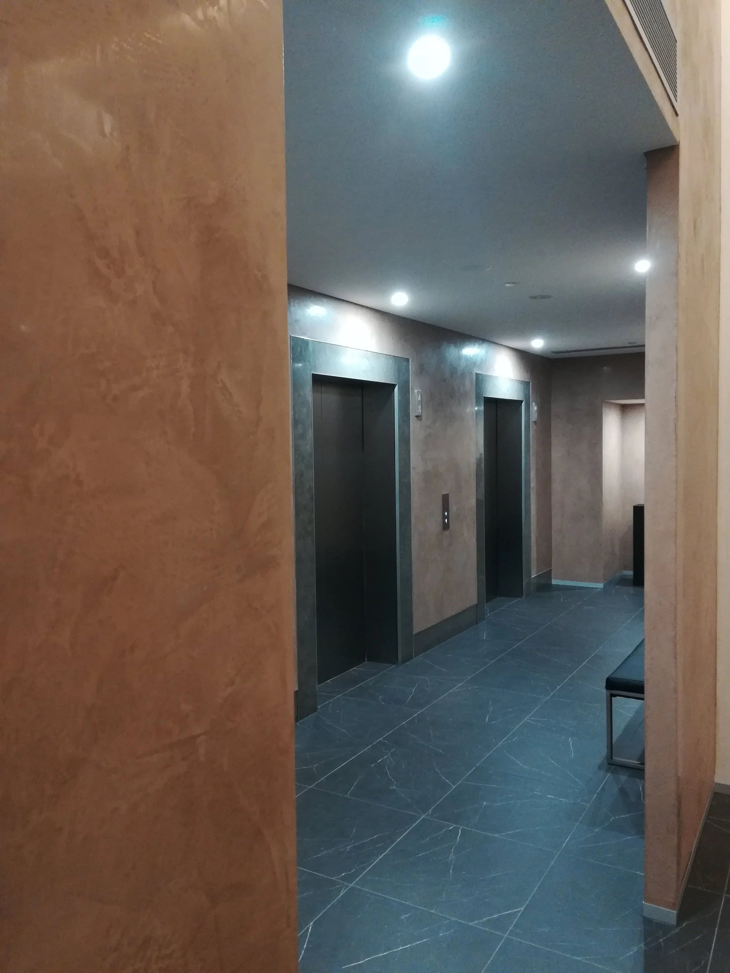 Two elevators with closed doors in a modern building hallway, brown textured walls, and dark tiled floor, with a bench on the right side.