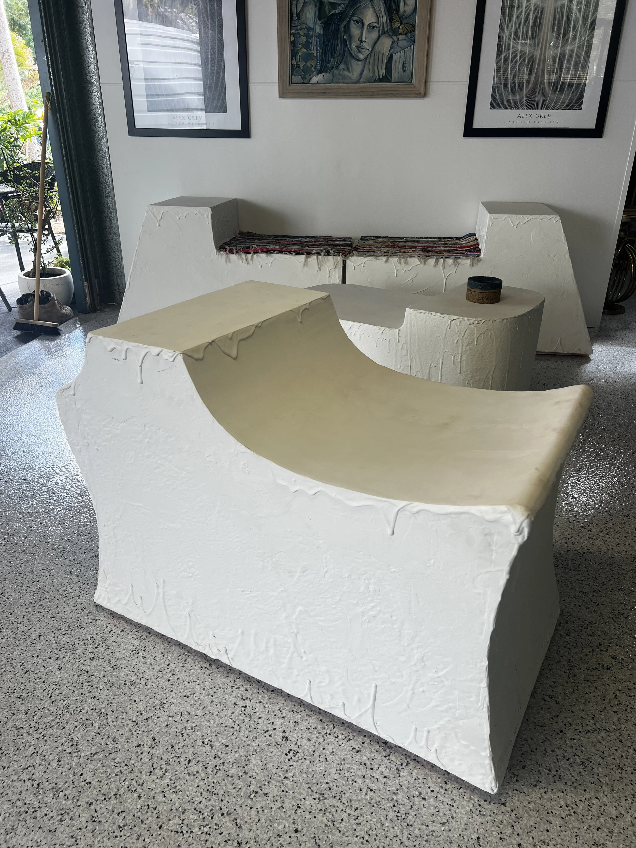 White sculptural furniture set consisting of a curved sofa and two matching block seats in a gallery or showroom. Artworks adorn the wall behind the furniture.