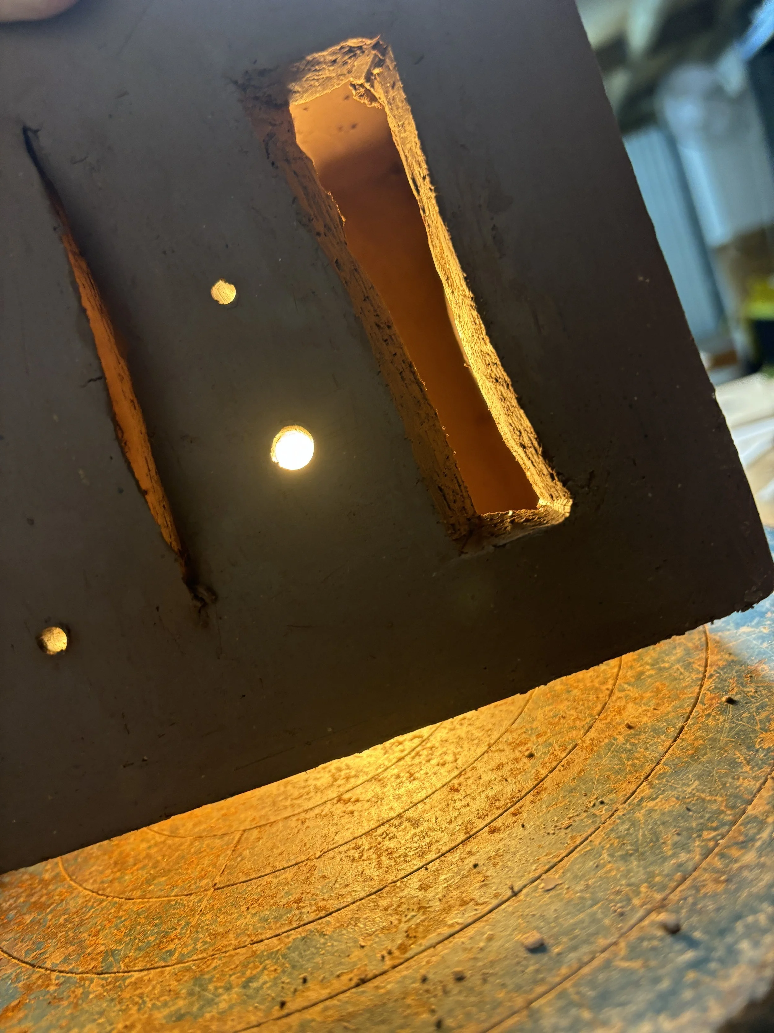 Close-up of a rusty metal piece with a rectangular hole and small round holes, with warm light shining through the openings.