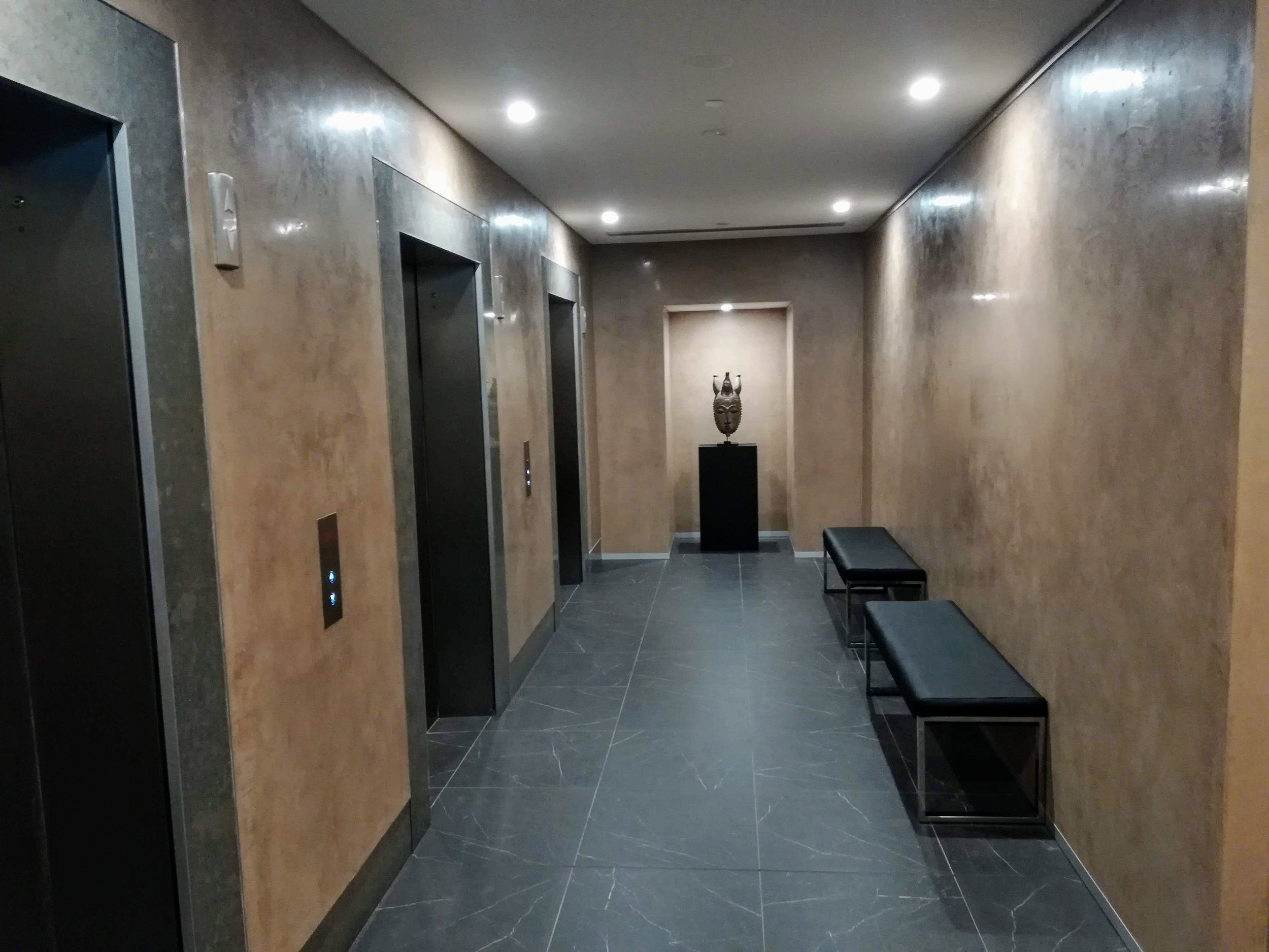 A hallway with two elevators, a sculpture on a pedestal at the end, and two black benches along the right wall.