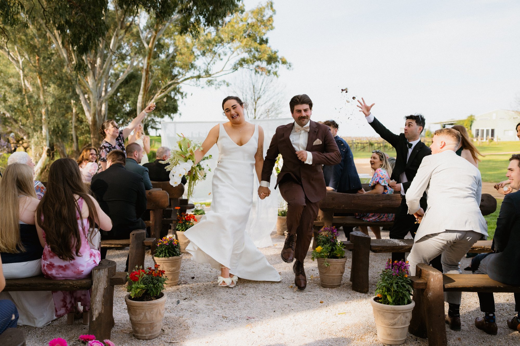 A newlywed couple walking down the aisle during an outdoor wedding ceremony, surrounded by friends and family, with some guests celebrating and throwing flower petals.
