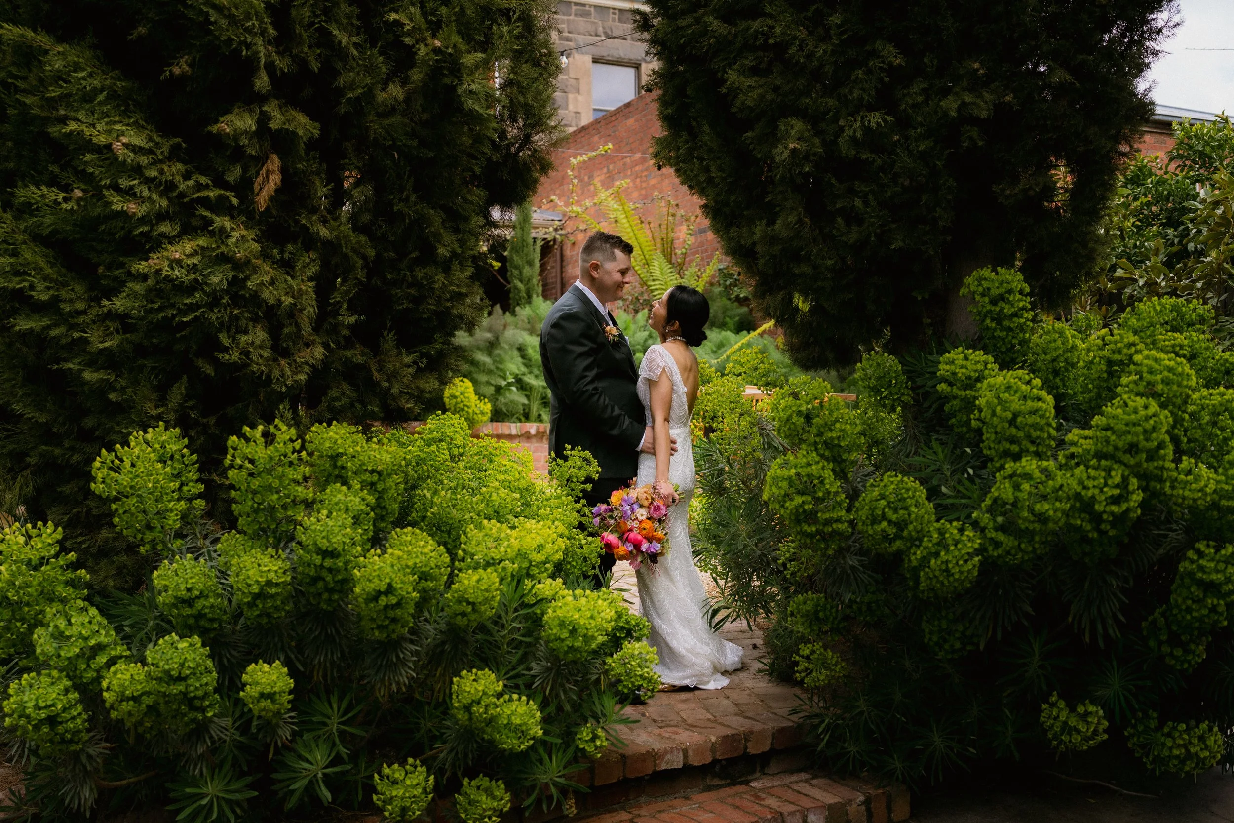 A bride and groom standing close together outdoors on a brick pathway, surrounded by lush green bushes and tall trees, sharing a tender moment. The bride holds a colorful bouquet of flowers.