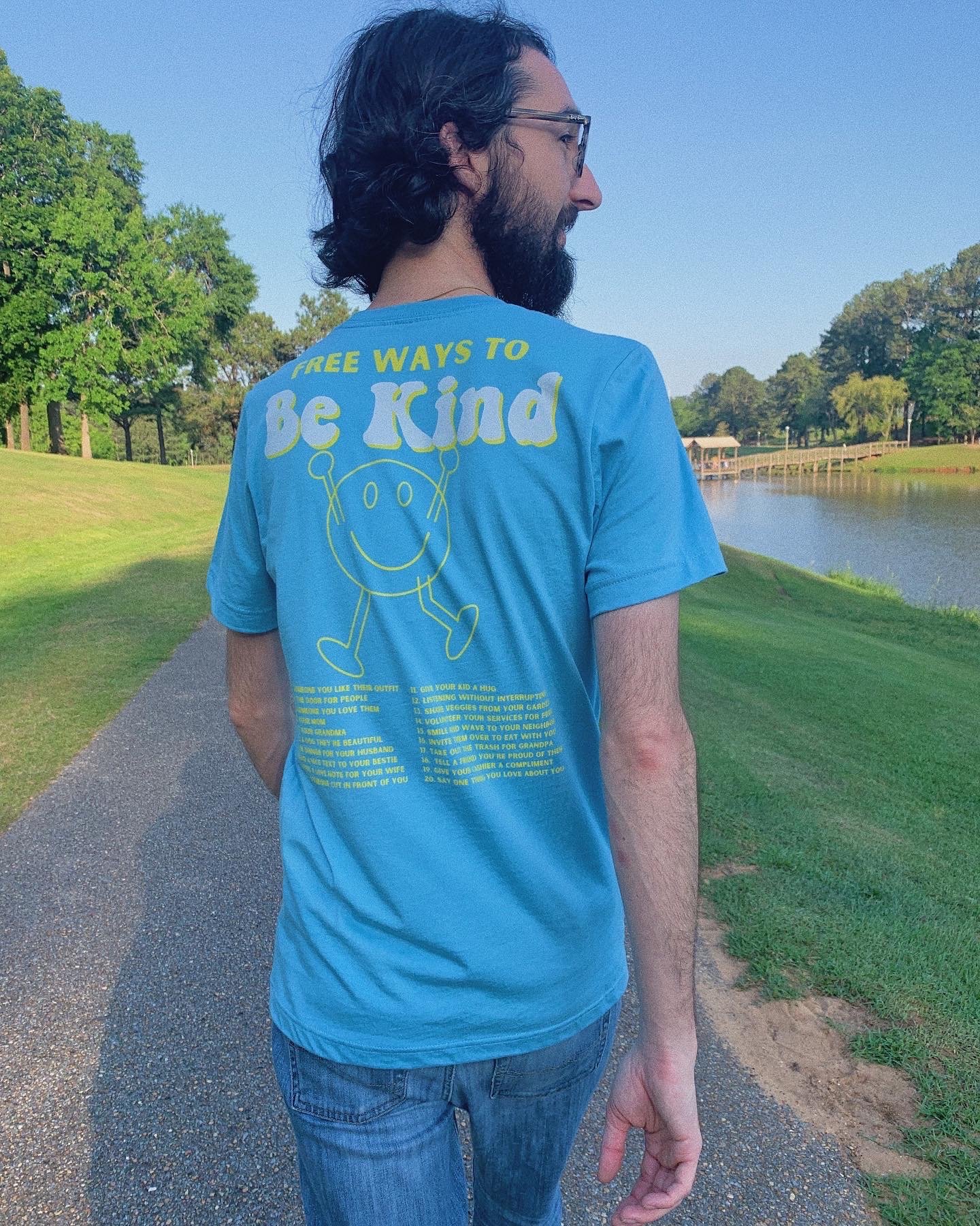 A young brunette man wearing the light blue go cozy tee. He is walking on a path thru the park and smiling. Be kind design