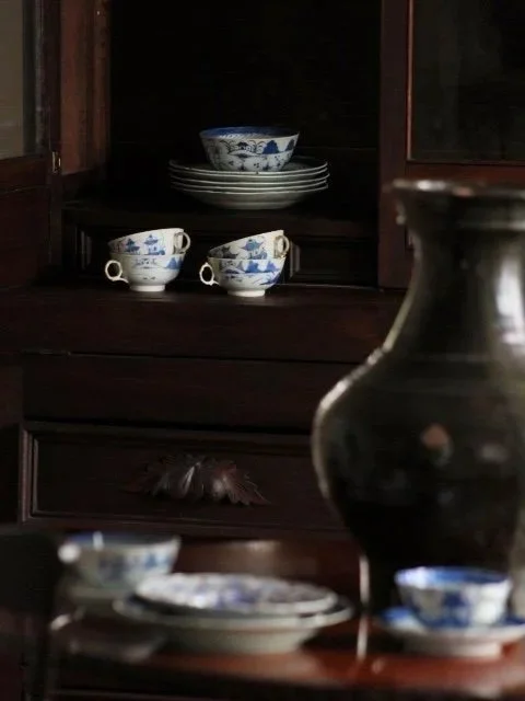 A dark wooden cabinet with blue and white patterned dishware, including bowls and teacups, and a large dark vase nearby.