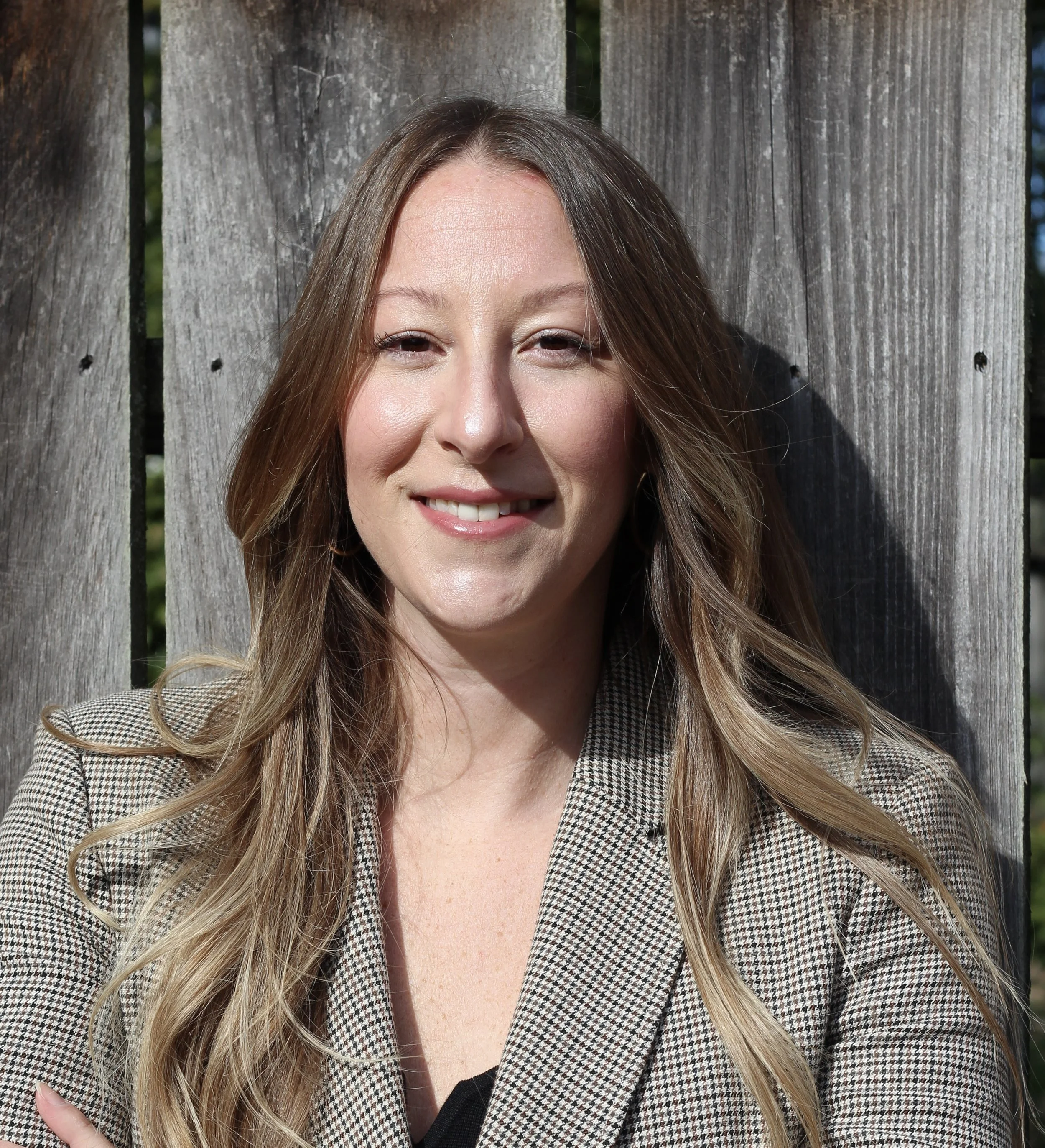 Close-up of a smiling woman with long wavy blonde hair, wearing a checkered blazer, standing outdoors against a wooden fence.