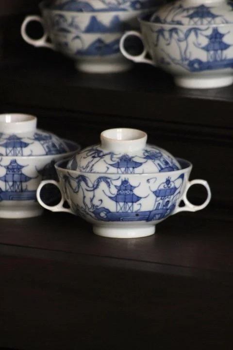 A set of blue and white patterned Chinese soup bowls with small handles, stacked inside each other, on a black shelf.