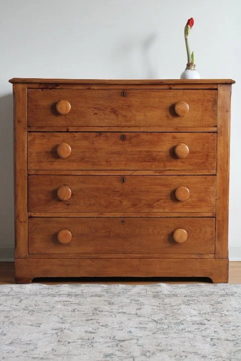 A wooden dresser with five drawers and round knobs, topped with a single red flower in a white vase, against a white wall.