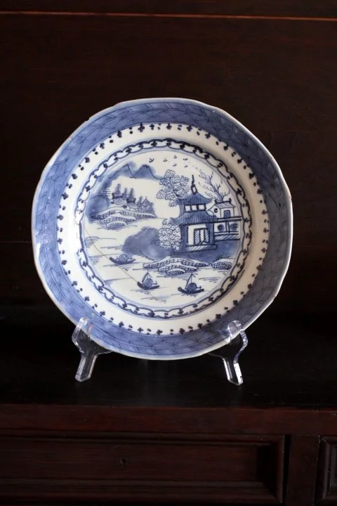 Blue and white patterned ceramic plate with a landscape scene, including a house, trees, mountains, and sailboats, displayed on a clear stand.