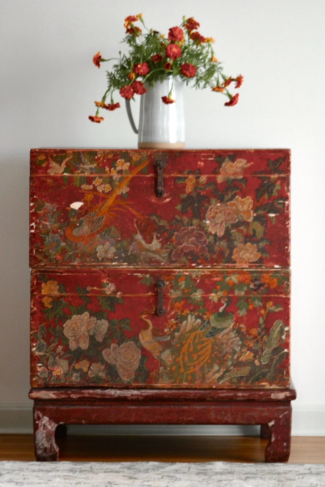 A tall, red, ornate wooden chest with floral and animal designs, placed against a plain white wall, with a white vase of orange flowers on top.