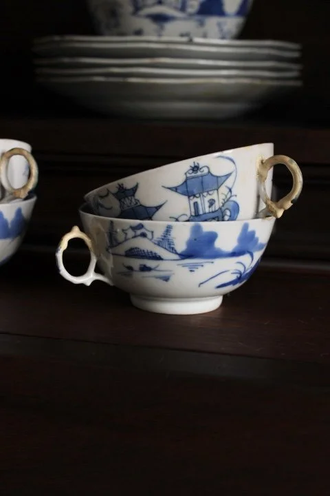 Stacked blue and white porcelain teacups with handles, featuring scenic and nautical designs, on a dark wooden surface.