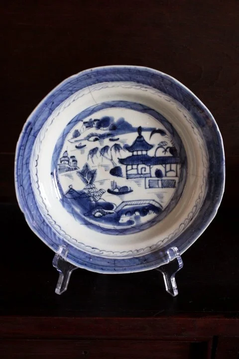 Blue and white porcelain bowl with traditional Asian landscape scene, including pagoda, trees, bridge, and boats, displayed on a clear stand.