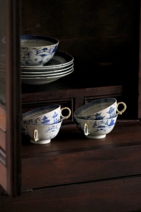 A set of blue and white porcelain tea cups with handles on a wooden shelf, with matching bowls in the background.