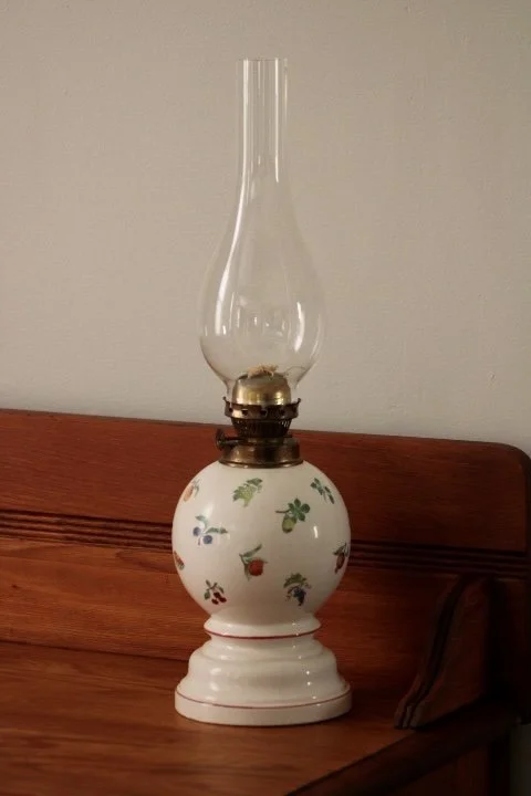 Decorative oil lamp with a floral patterned ceramic base and a glass chimney, placed on a wooden surface against a plain wall.