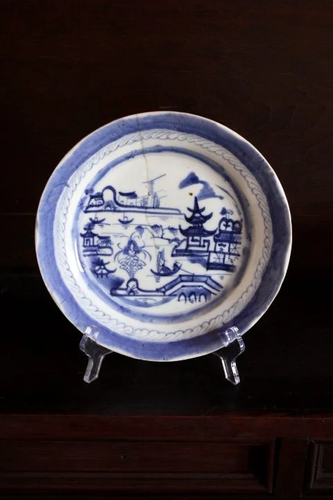 Blue and white decorative plate with an Asian-inspired landscape scene, including buildings, trees, water, and a sailboat, displayed on a clear stand.