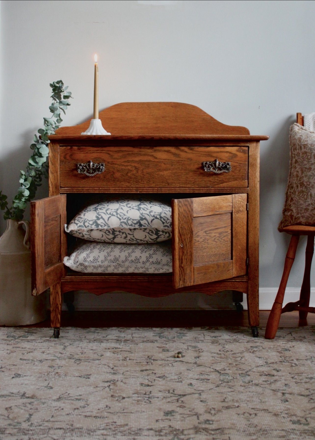 A wooden cabinet with open doors revealing two drawers and two shelves with fabric pillows inside. A lit candle in a white holder and a green leafy branch are on top of the cabinet. To the right, a chair with patterned fabric and wooden legs is visib