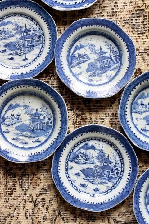 A collection of blue and white porcelain plates with scenic landscapes, buildings, trees, and water, arranged on a textured fabric surface.