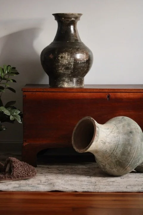 Two antique clay pots, one upright on a wooden surface and the other tilted on the floor, with a plant and a wooden piece of furniture in the background.
