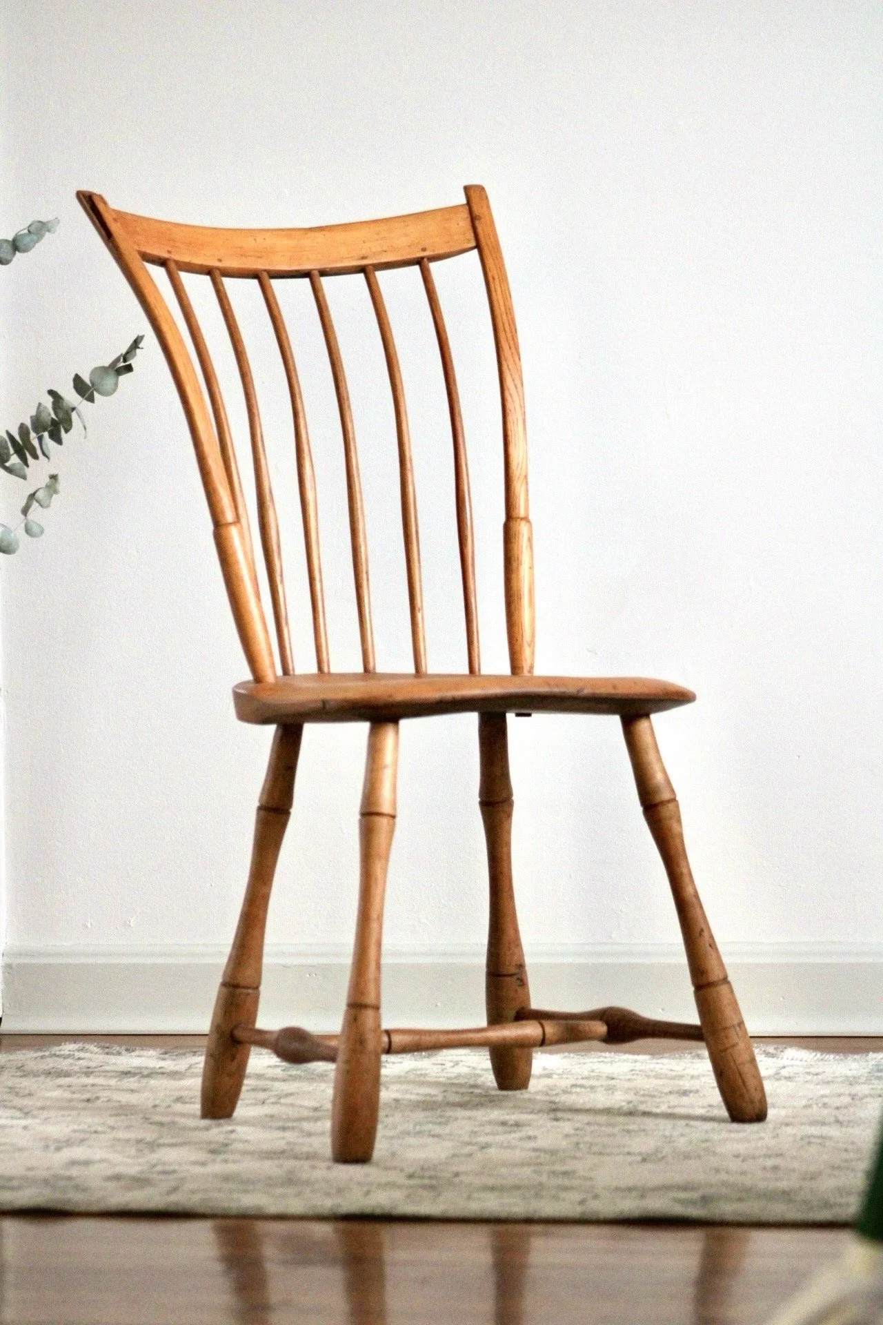 A wooden chair with a curved backrest and vertical spindles, standing against a plain white wall, with a plant partially visible on the left and a textured rug on the floor.