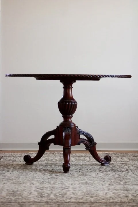 A vintage wooden pedestal table with a round top, placed on a patterned rug against a plain white wall.