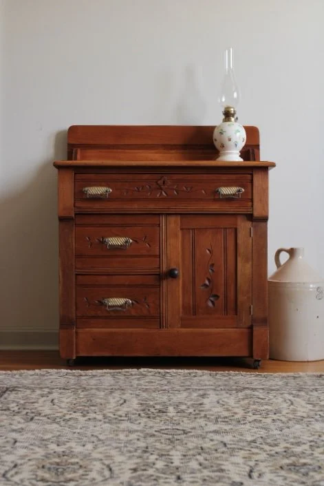 A wooden dresser with drawers and a door, topped with a decorative lamp. A large ceramic jug is on the floor to the right of the dresser. A patterned rug covers the floor in front of the dresser.
