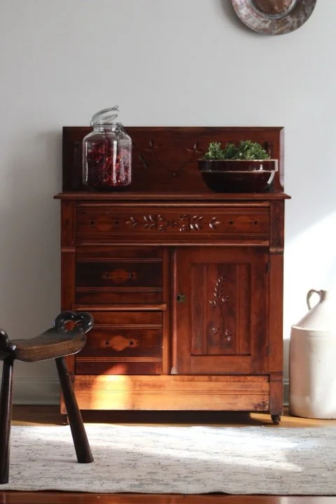 A wooden cabinet with decorative carvings, topped with a glass jar filled with red decorative items and a large bowl of green leafy salad, against a light-colored wall with a wall hanging above it.