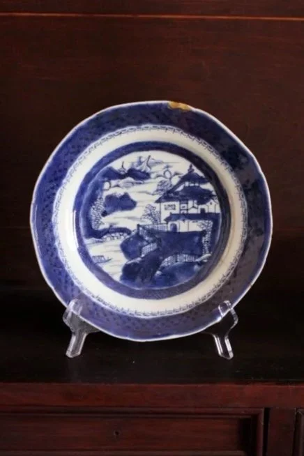 A blue and white decorative plate with a landscape scene, displayed on a clear stand on a dark wooden surface.