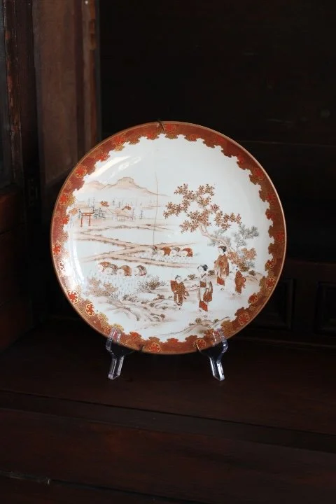 Decorative ceramic plate with a rural winter landscape painting, featuring people, animals, and trees in sepia tones, displayed on a stand.