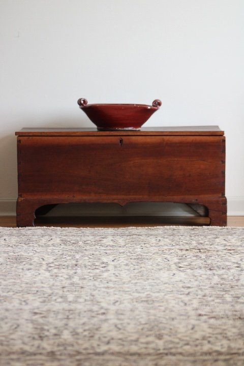 A wooden bench with a red decorative bowl on top, against a plain white wall, on a light-colored rug.
