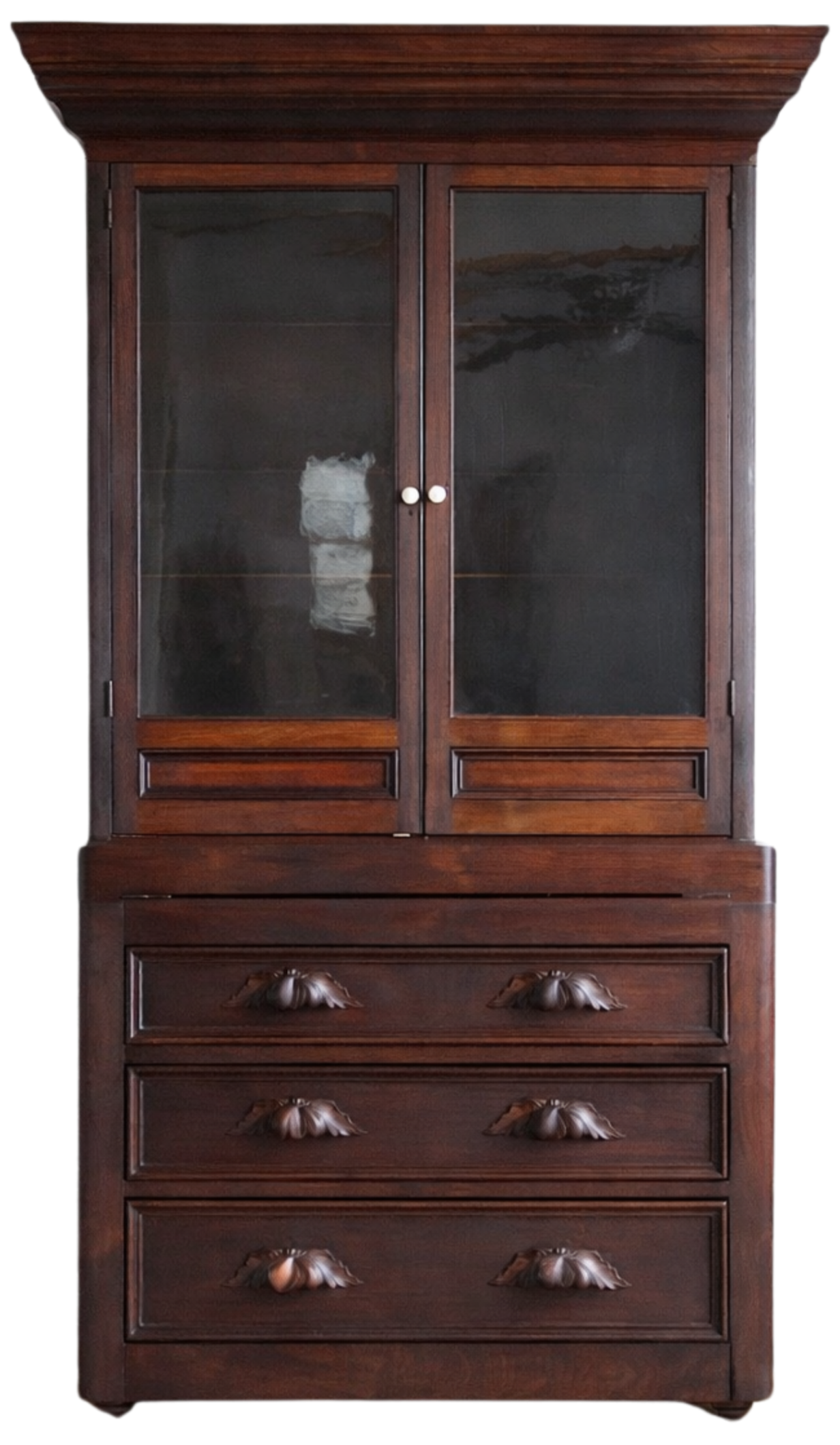 A tall, antique wooden cabinet with glass doors on the top section and multiple drawers below, featuring ornate carved handles.