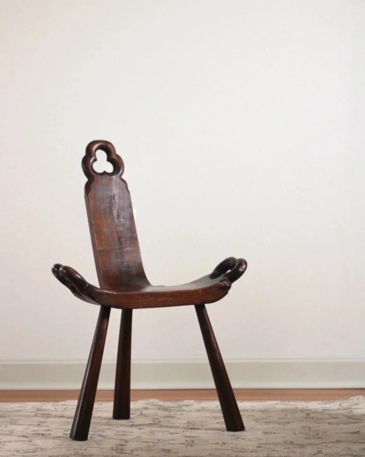 A wooden chair with a tall, decorative backrest shaped like a keyhole, placed against a plain, light-colored wall and on a patterned area rug.