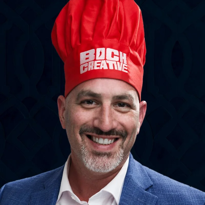 A man wearing a blue suit and a white shirt, smiling, with a red chef hat that says "BOCH CREATIVE" on it.