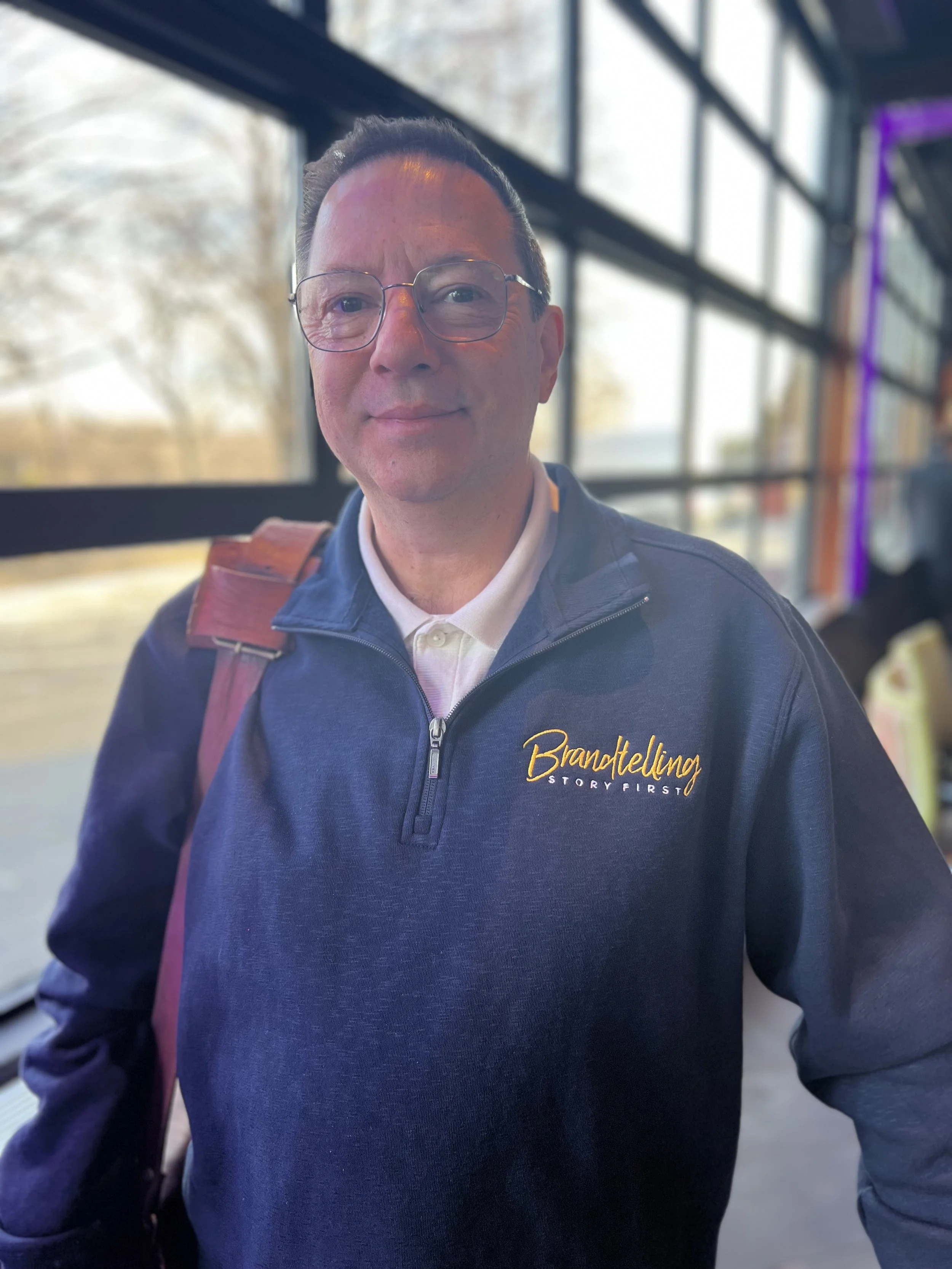 A man with glasses wears a navy blue jacket with the word 'Brandtelling' embroidered on it. He stands indoors near large windows with a blurred outdoor background.