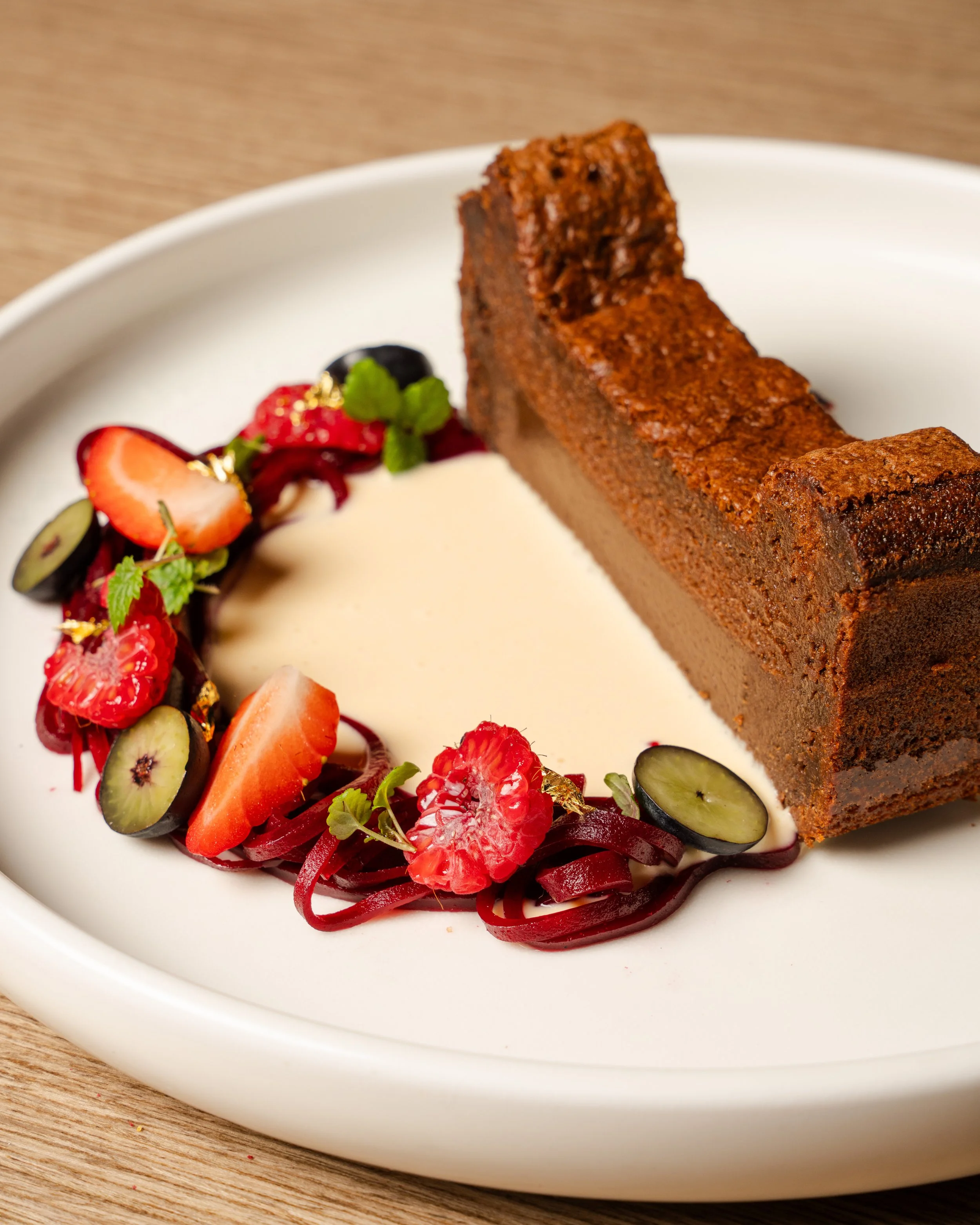 A slice of chocolate cake with a dense, rich texture on a white plate, garnished with fresh berries, mint leaves, and edible gold flakes.