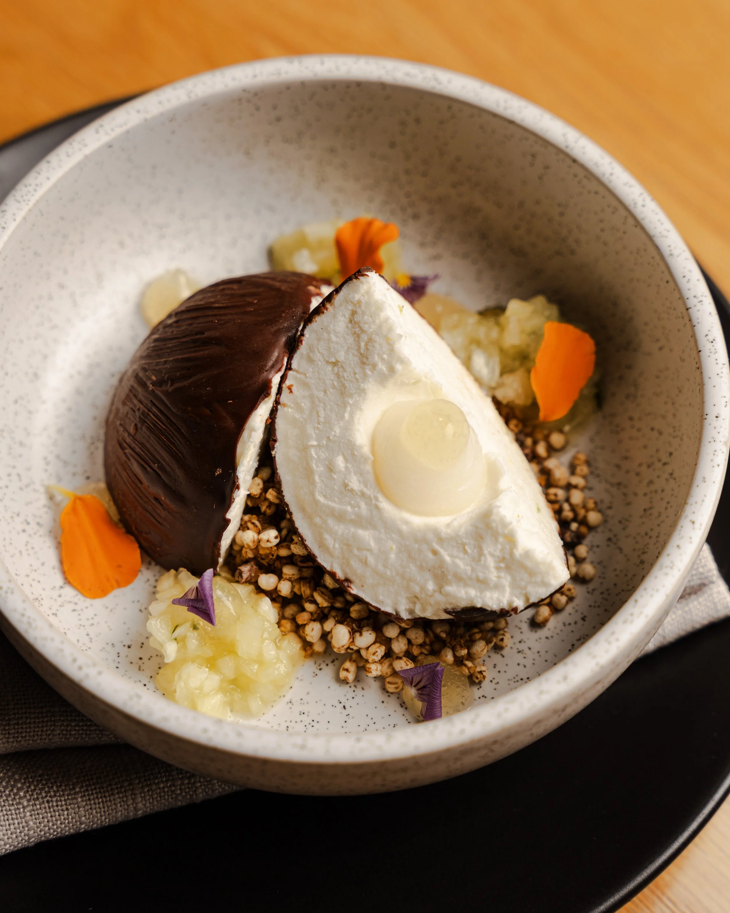 A slice of cheesecake with a dark chocolate topping, garnished with edible flowers, surrounded by quinoa or millet grains, and small dollops of fruit compote, in a speckled ceramic bowl.