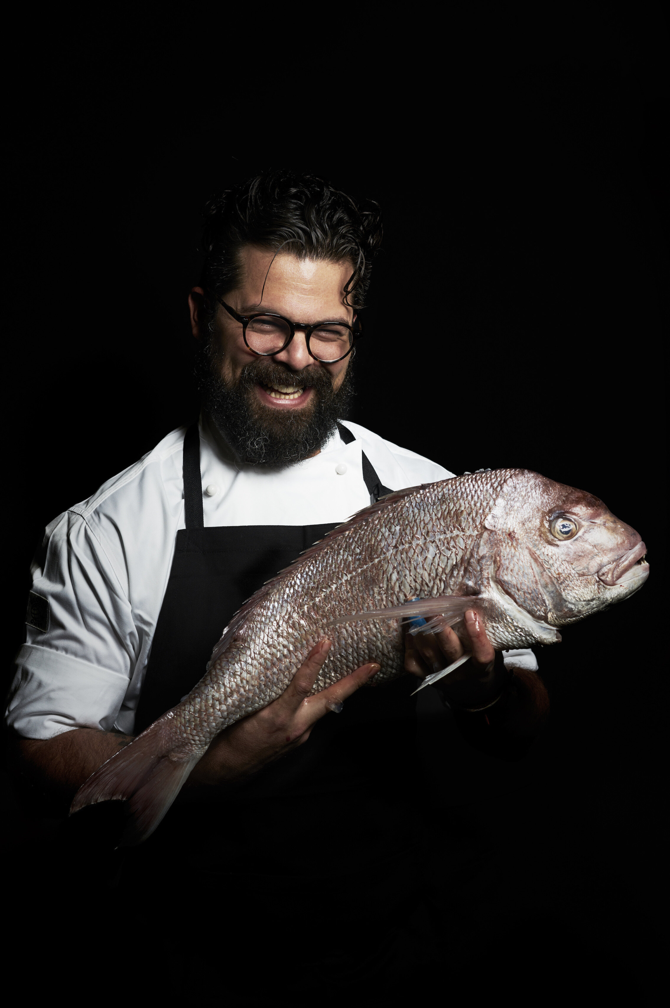 A man with curly hair, glasses, and a beard is smiling and holding a large fish, wearing a white chef's coat and black apron against a black background.