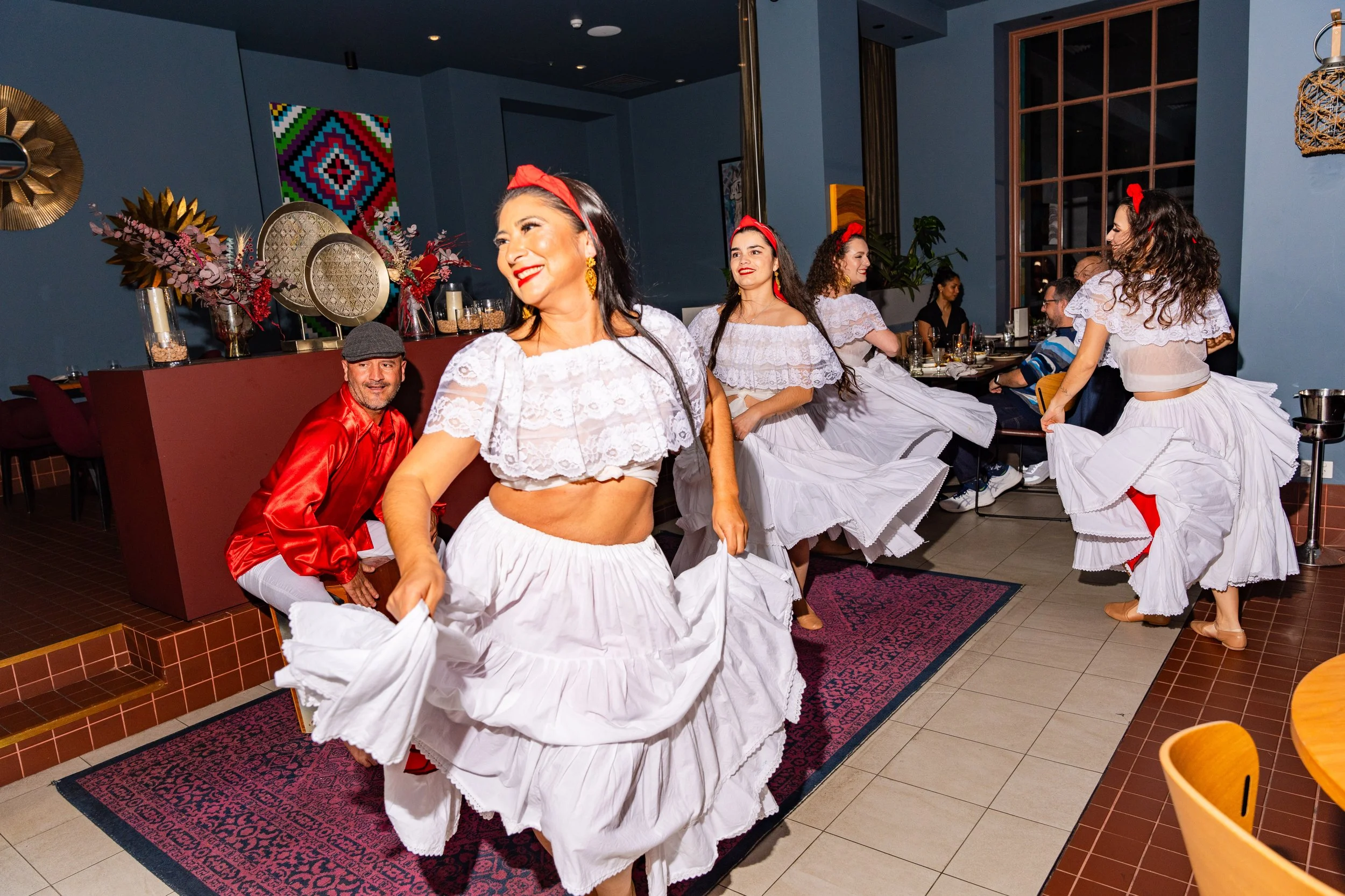 Group of women dressed in traditional folkloric white dresses with red accents, dancing and smiling in a restaurant with dark blue walls and colorful decorations.