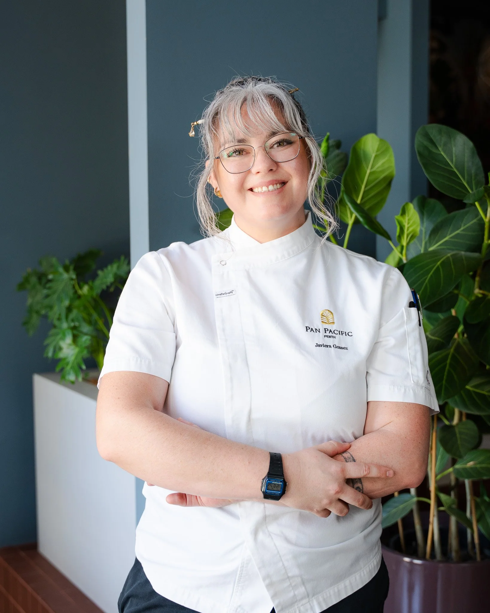 A woman with gray hair, glasses, and a chef's uniform smiling with arms crossed, standing indoors with green houseplants behind her.