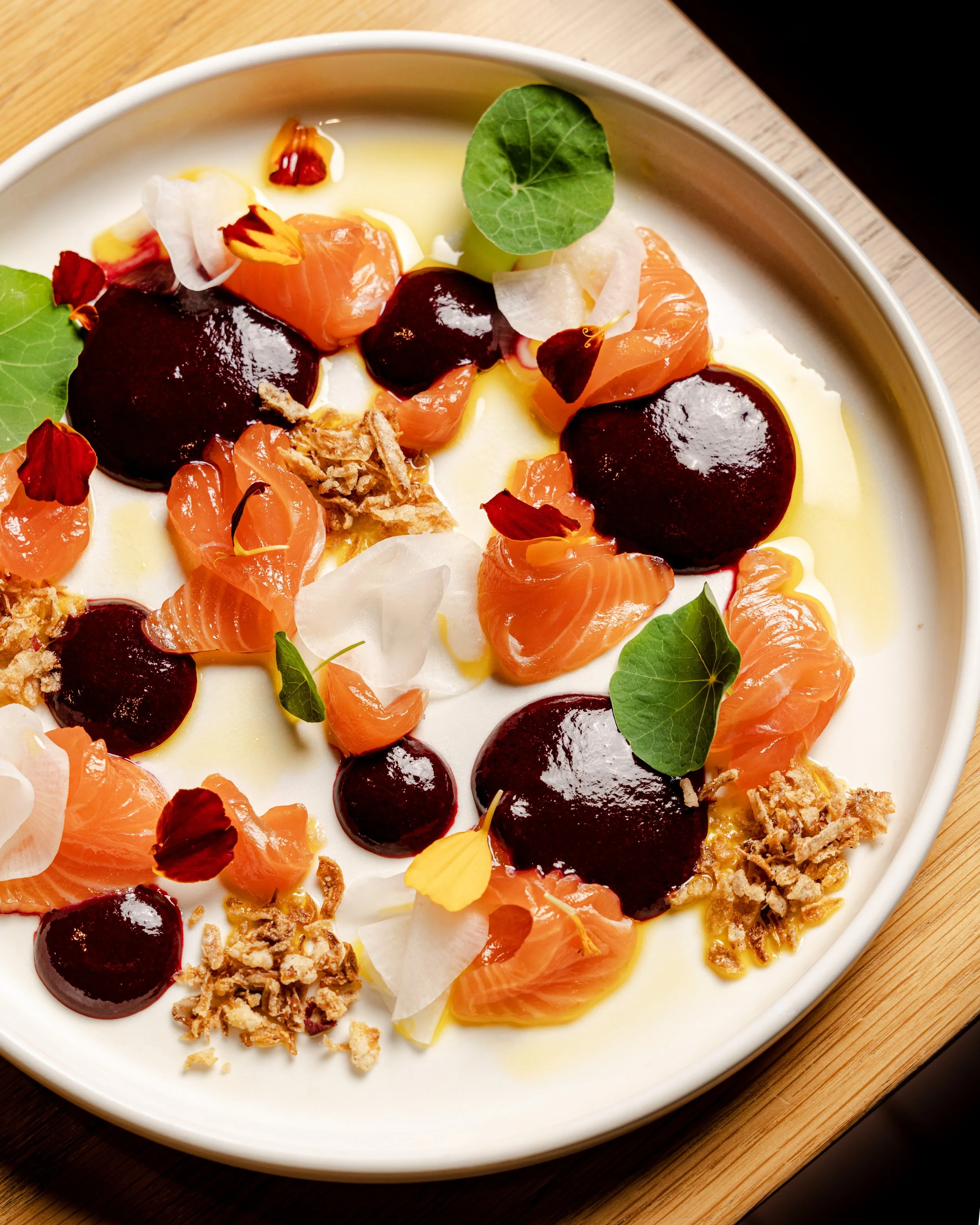 A dessert dish with dollops of dark berry sauce, orange salmon roes, edible flowers, and crunchy garnishes on a white plate.