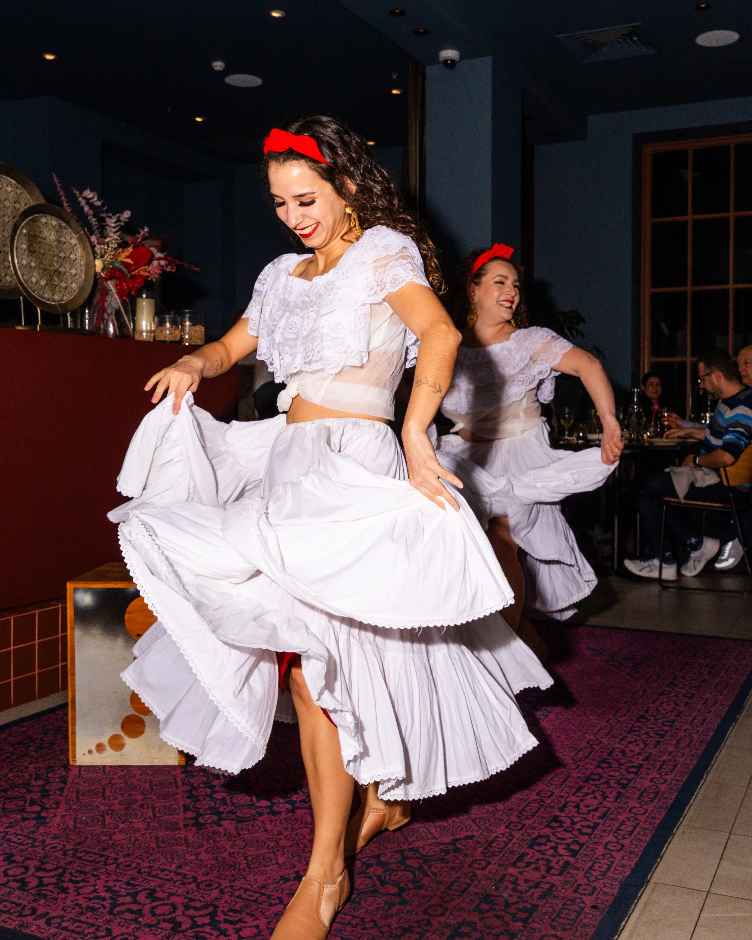 Two women dressed in traditional Mexican folkloric dresses dance indoors on a dark carpeted floor, with one woman visibly smiling and raising her leg.